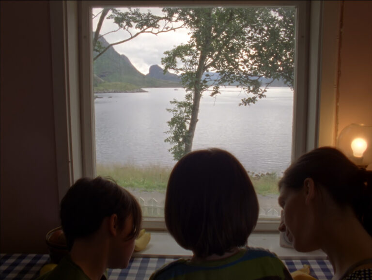 Two children and a woman and pictured in front of a window with their backs turned to the camera. They appear in from of a table with a blue and white gingham tablecloth. The table faces a window which looks onto a lake with a tree in front and rolling hills in the distance.