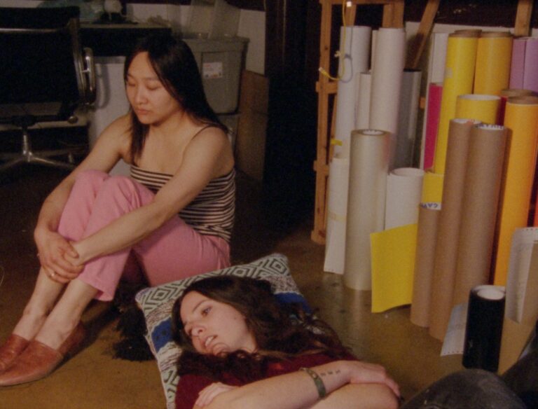 two women are pictured; one is sitting with her legs folded with her joined hands resting on her shins. She has long dark hair and is wearing a black and white striped top with pink trousers and tan slip-on shoes. The second woman is seen from the waist up; she is lying on the floor with her arms crossed. She has long dark hair and is wearing a dark top. Both women are facing away to their right. Beside them we see a collection of brown, white, yellow, pink and black paper which is rolled up and placed upright.