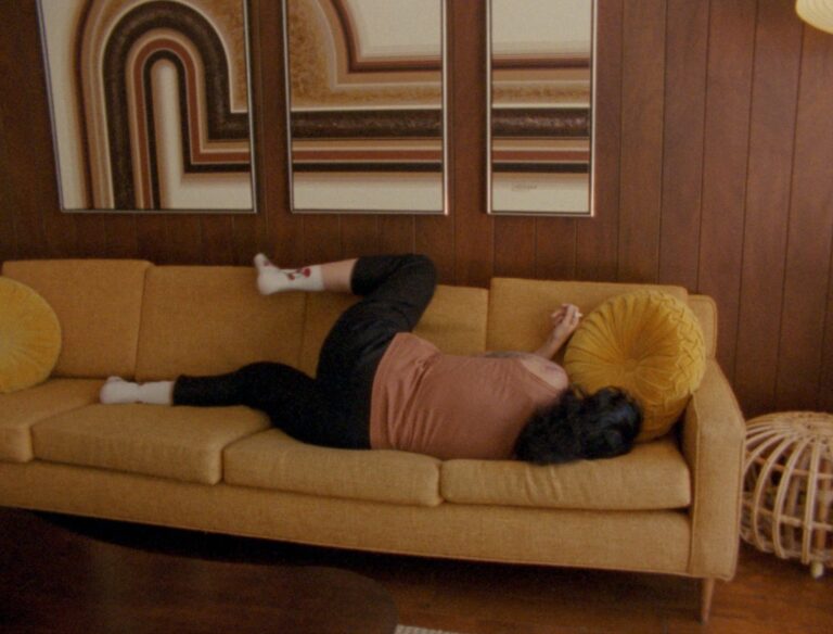 A person lies on a mustard sofa facing away from the camera. They are wearing dark trousers, a peach coloured sleeveless top and white socks. They have dark hair and their left leg is raised up and rests along the top of the sofa. The room is panelled in dark wood with a triptych of 70s style linear paintings on the wall behind the sofa.