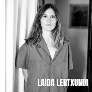 The artist Laida Lertxundi is pictured in a black and white shot. She is wearing a short sleeved shirt and looks directly at the camera. She has long hair reaching past her shoulders and her arms are crossed behind her back. She stands beside a curtain and a framed painting is seen in the distance.