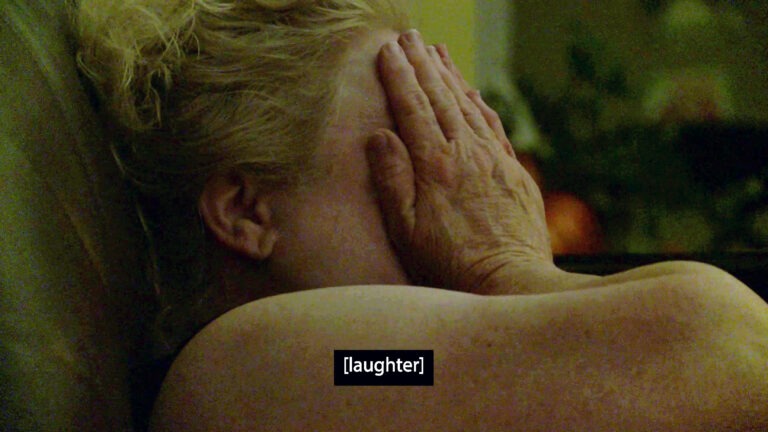 In a dimly lit room, a white woman with blonde hair covers her face with her hands. Her eyes appear to be closed tightly; it's unclear if she's laughing or crying. At the bottom of the screen, an open caption reads: 