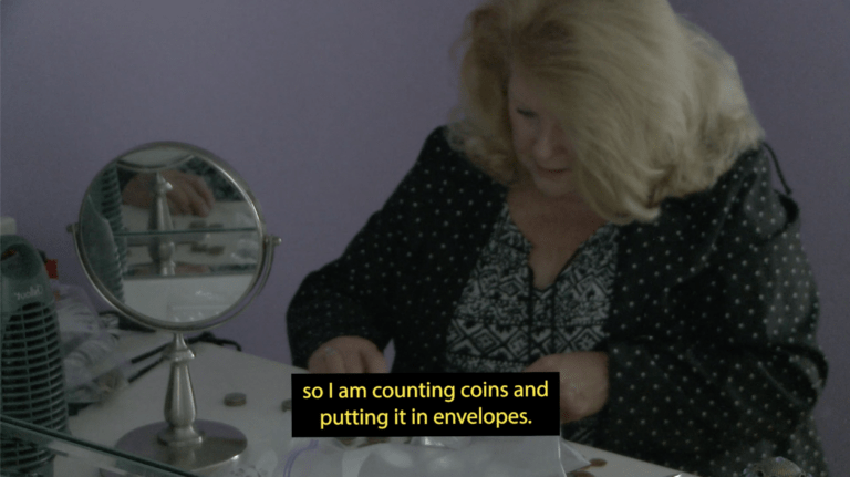 A white woman with shoulder-length blonde hair covering one eye sits, looking down, wearing a polka-dotted raincoat. A hand-held mirror sets on a counter next to her, reflecting her hand and stacks of coins. A caption appears on top of her hands that reads “so I am counting coins and putting in envelopes.”