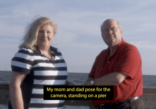 A white woman with shoulder-length blonde hair stands next to a bald man wearing glasses, in front of an ocean and a cloudy blue sky. They both stare at the camera. A caption at the bottom of the image reads: "My mom and dad pose for the camera, standing on a pier".