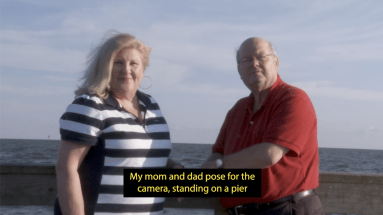 A white woman with shoulder-length blonde hair stands next to a bald man wearing glasses, in front of an ocean and a cloudy blue sky. They both stare at the camera. A caption at the bottom of the image reads: "My mom and dad pose for the camera, standing on a pier".