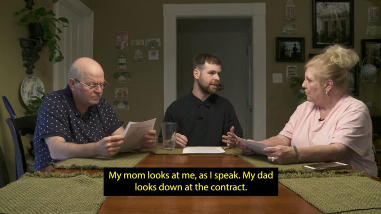 Three white people sit at a table together, each with a set of white papers in front of them. The figure on the far left is a bald man in his 60s, who reads the papers intently. The figure in the center is a person in their late 20s, who looks to a woman in her 60s with blonde hair, who holds the papers in her hands and looks back at them. A caption appears over the image that reads: “My mom looks at me, as I speak. My dad looks down at the contract.”