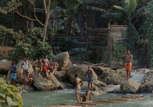 Ten young black men are gathered beside a rivers edge. Some are seated on rocks, some are standing and two are on a wooden raft. Behind them appears a riverside dwelling.